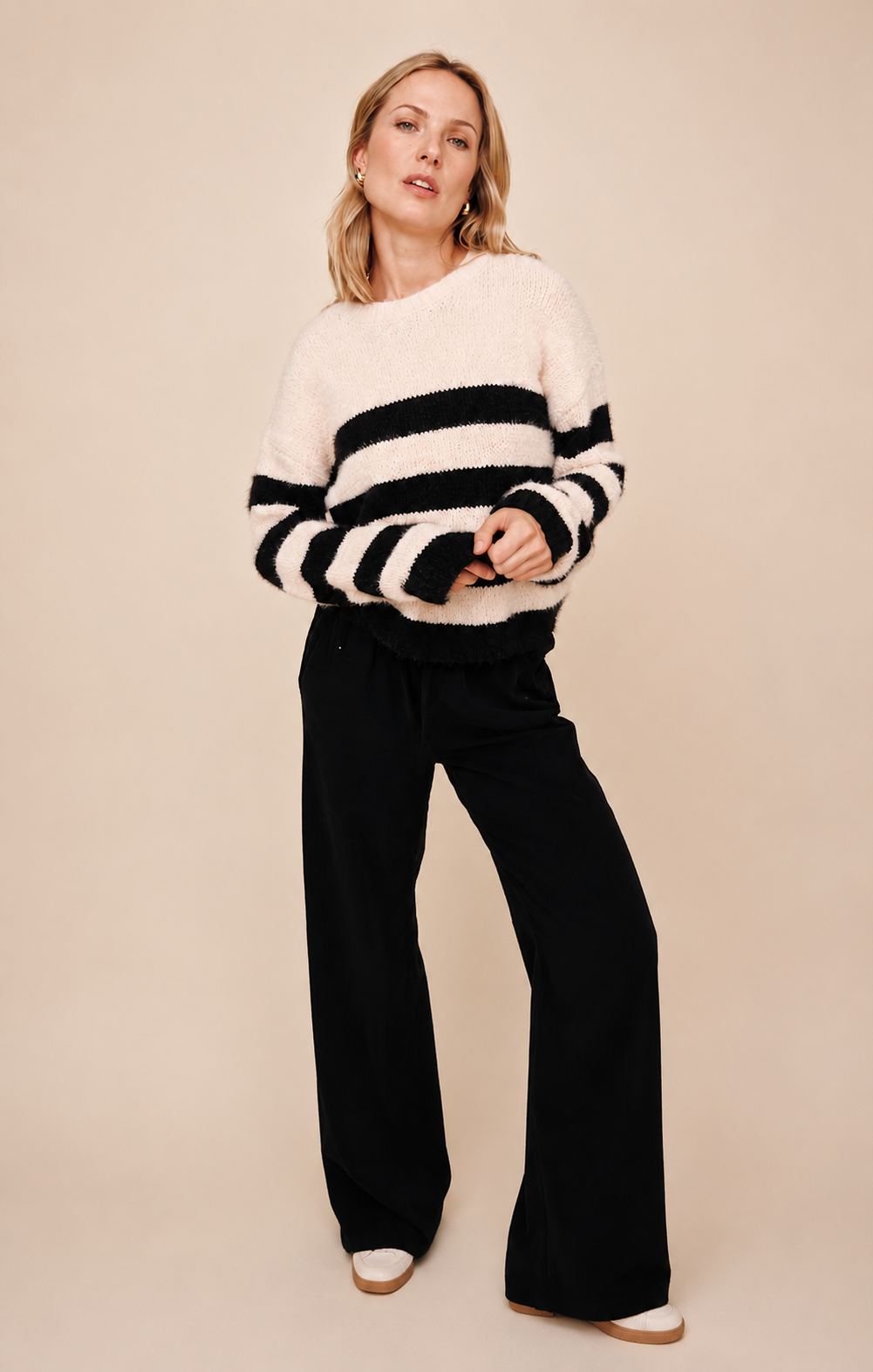 A woman modeling a fuzzy knit sweater with horizontal black and cream stripes, paired with black wide-leg pants, against a solid beige studio background.