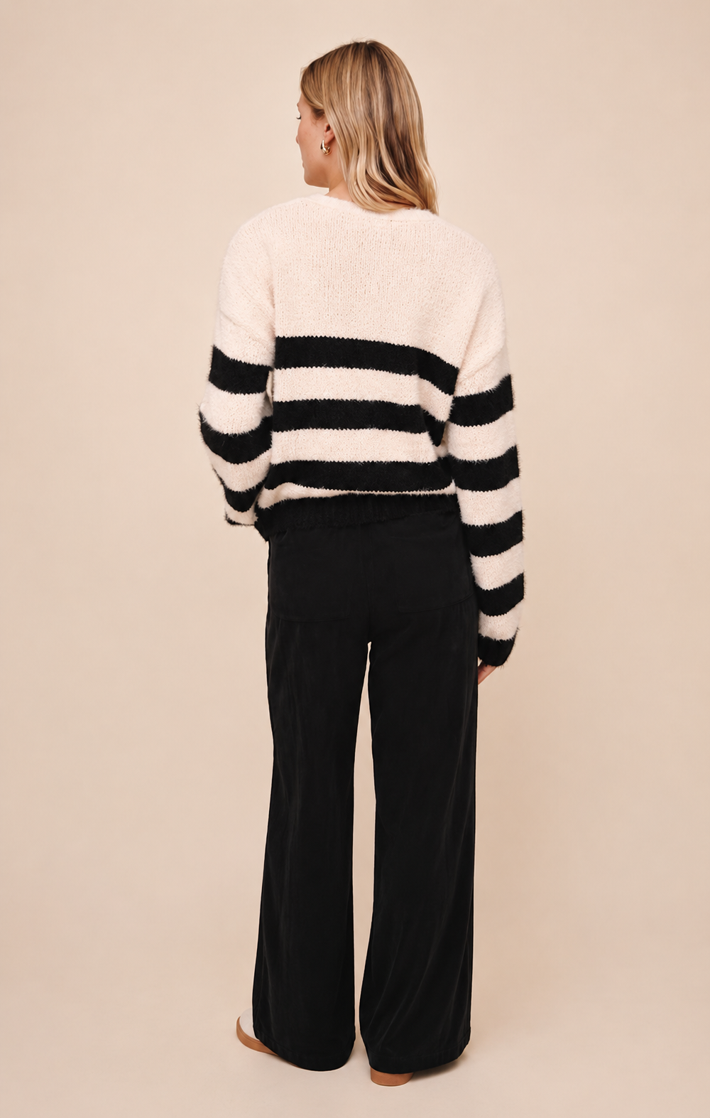 A woman shown from the back wearing a fuzzy knit sweater with horizontal black and off-white stripes and black wide-leg pants, standing against a plain beige studio background.