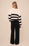 A woman shown from the back wearing a fuzzy knit sweater with horizontal black and off-white stripes and black wide-leg pants, standing against a plain beige studio background.