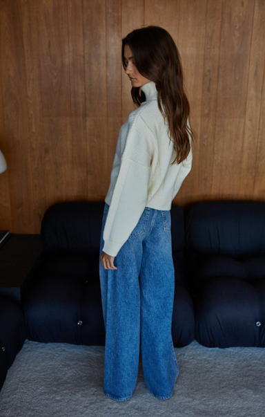 Woman wearing a cream sweater and blue jeans standing in a room with wooden walls and dark furniture.