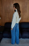 Woman wearing a cream sweater and blue jeans standing in a room with wooden walls and dark furniture.