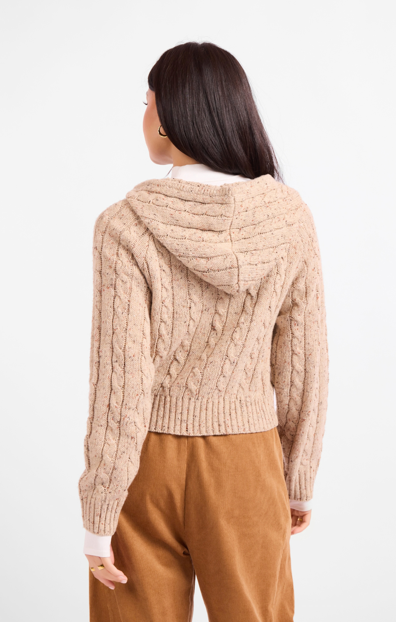 Woman wearing a beige cable knit hoodie and brown pants on a white background