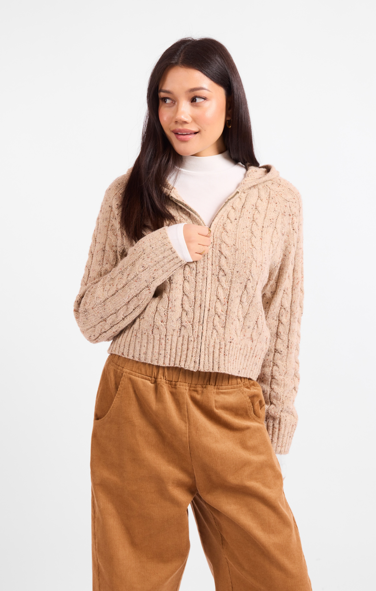 Woman wearing a beige cable knit sweater and brown pants on a white background