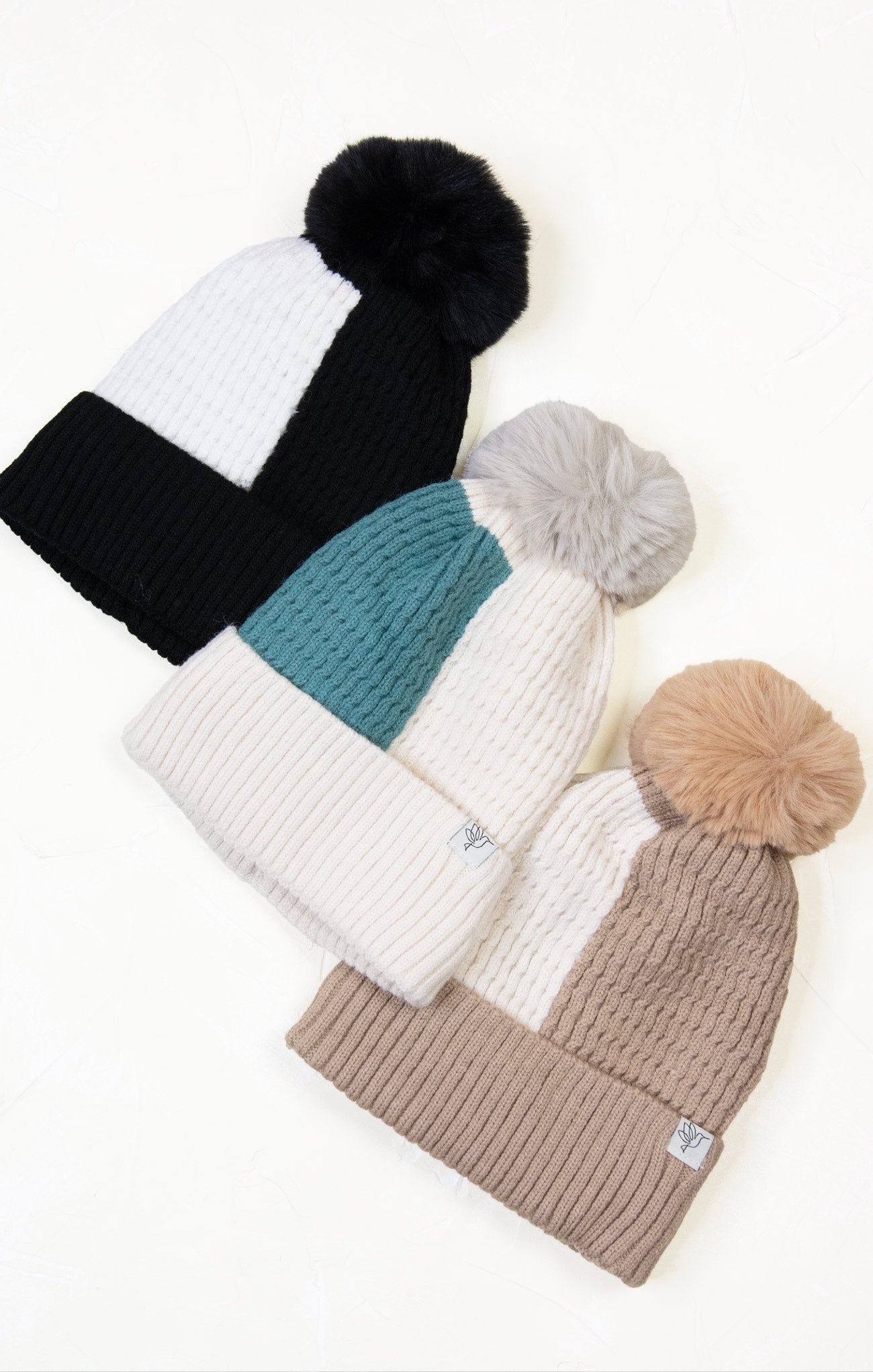 Four knit beanie hats with pom-poms in black, white, teal, and beige on a light background.