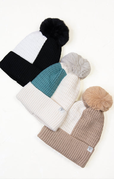 Four knit beanie hats with pom-poms in black, white, teal, and beige on a light background.