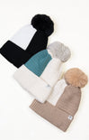 Four knit beanie hats with pom-poms in black, white, teal, and beige on a light background.