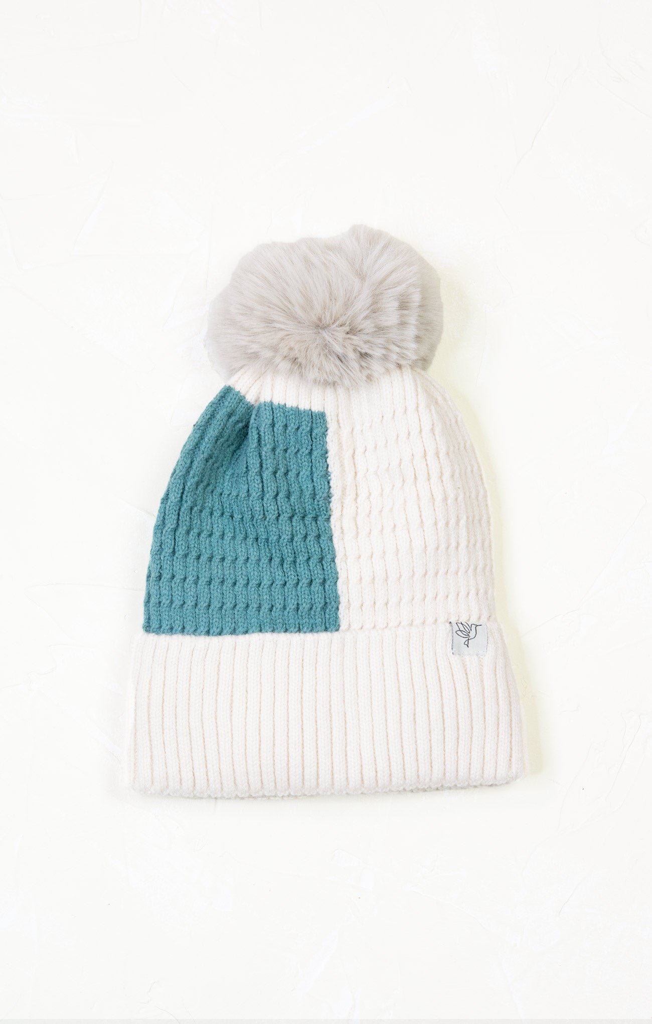 White knit beanie with a teal section and gray pom-pom on a white background