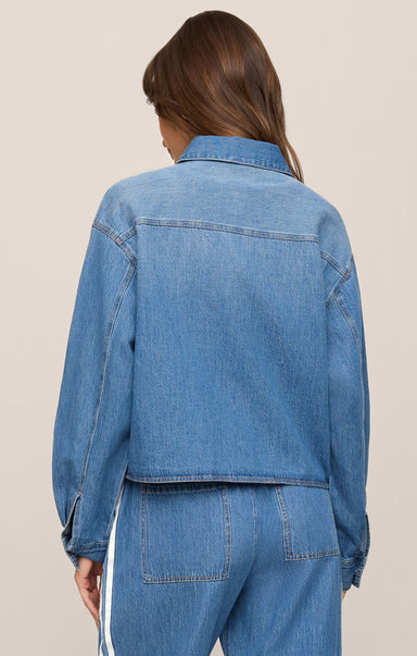 A woman shown from the back wearing a cropped blue denim jacket and matching blue denim pants against a plain beige studio background.