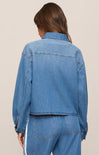 A woman shown from the back wearing a cropped blue denim jacket and matching blue denim pants against a plain beige studio background.