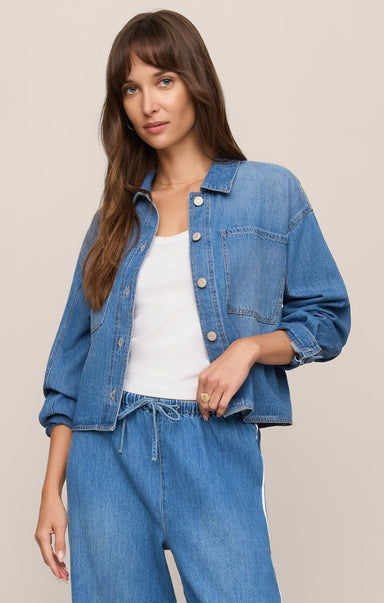 A woman modeling a cropped blue denim jacket over a white tank top and matching blue denim drawstring pants, photographed against a plain beige studio background.