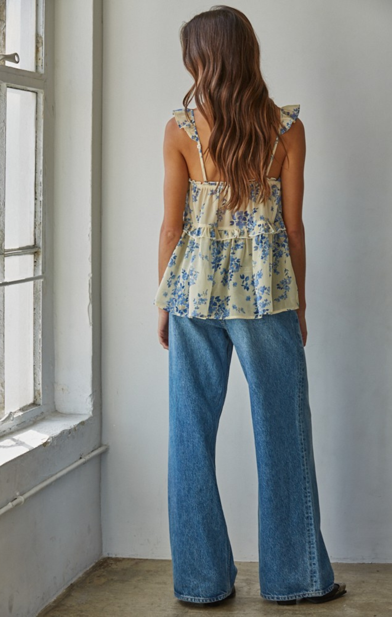 Woman wearing a floral top and blue jeans standing in a room with a window.