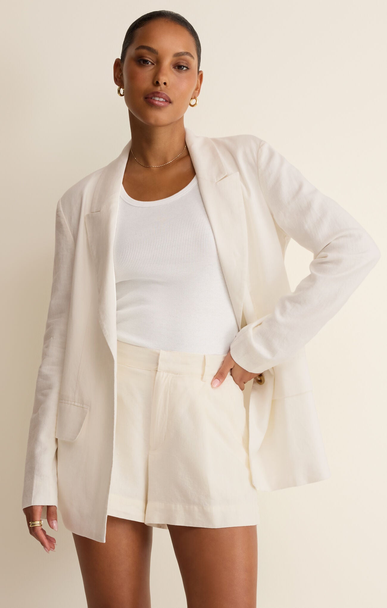 Woman wearing a white blazer over a white top and cream shorts on a beige background
