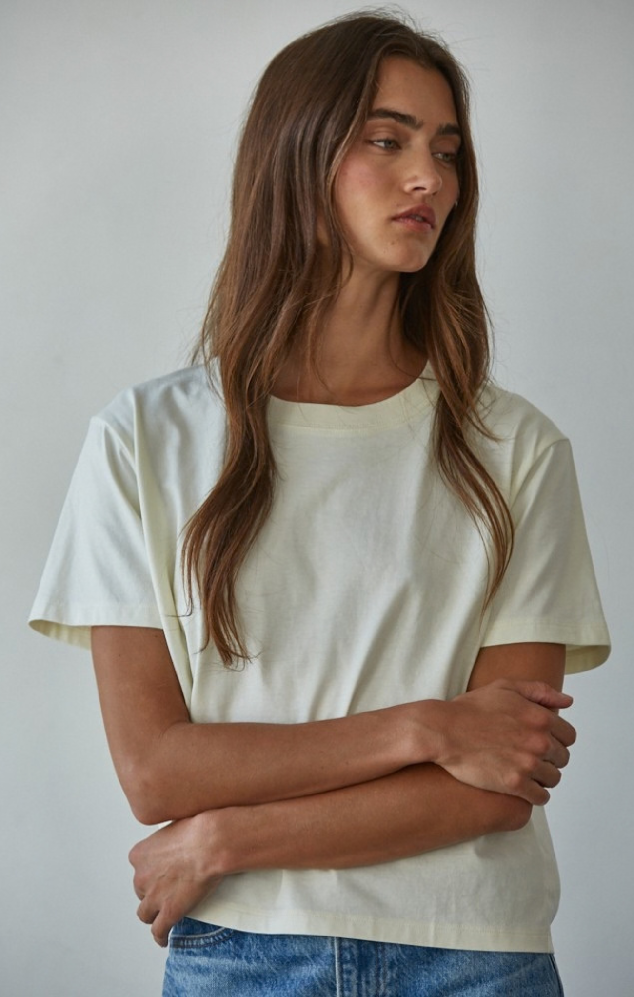 A woman with long brown hair wearing a cream-colored short-sleeve crewneck t-shirt and blue jeans, standing against a plain light gray background.