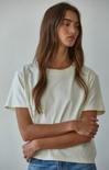 A woman with long brown hair wearing a cream-colored short-sleeve crewneck t-shirt and blue jeans, standing against a plain light gray background.
