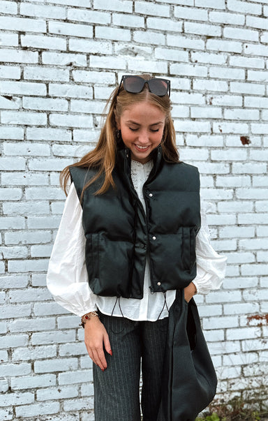 Woman wearing a black puffer vest over a white shirt against a light blue brick wall.