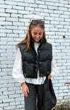 Woman wearing a black puffer vest over a white shirt against a light blue brick wall.