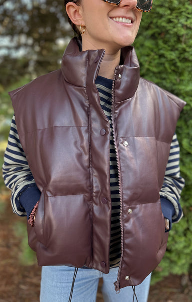 Person wearing a brown puffer vest with a striped shirt and sunglasses, standing outdoors.