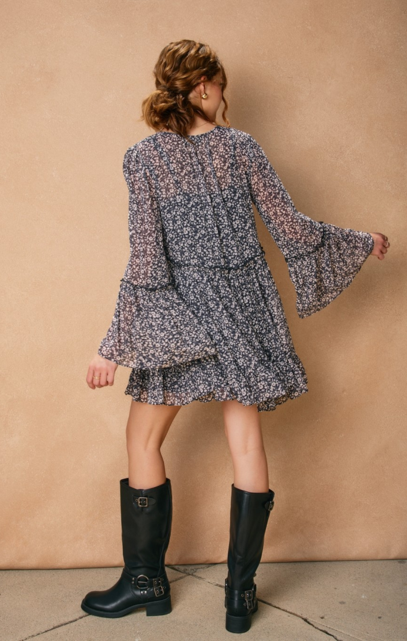 Person wearing a floral dress with bell sleeves and black rain boots against a beige wall.