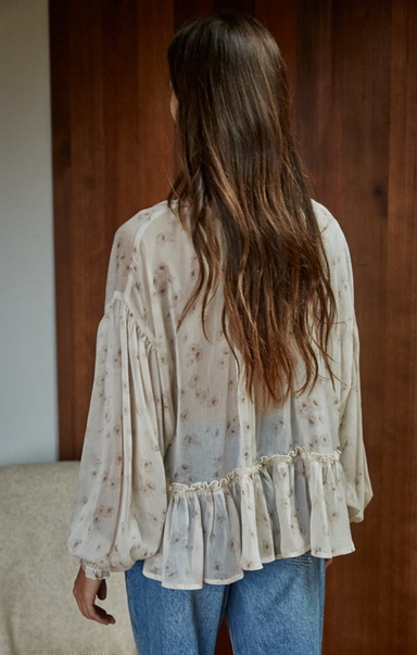 Woman wearing a light blouse with long sleeves and blue jeans, standing against a wooden wall.