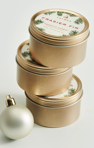 Stacked gold travel tins with a Christmas candle on top, labeled Frasier Fir, alongside a white ornament.