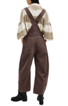 Person wearing brown overalls and a patterned shirt on a white background