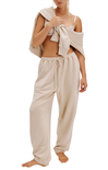 Woman wearing beige lounge pants and a matching top with a towel draped over her shoulders on a white background.