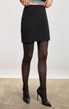 A model wearing a black A-line mini skirt, sheer black tights, and black patent high heels, standing against a neutral studio backdrop.