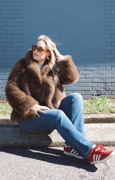 Person wearing a brown fur coat, blue jeans, and red Adidas sneakers sitting against a blue brick wall.