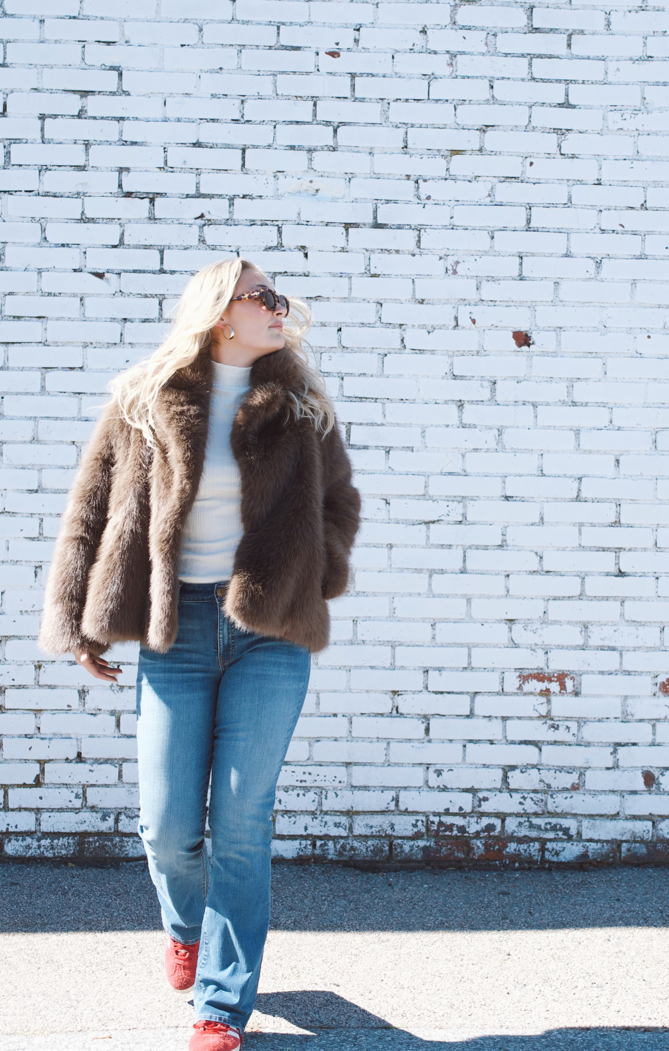 Person wearing a brown fur coat and jeans standing against a white brick wall.