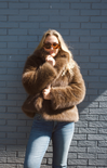 Person wearing a brown fur coat and sunglasses against a gray brick wall