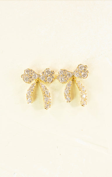 Gold bow-shaped earrings with clear gemstones on a light beige background