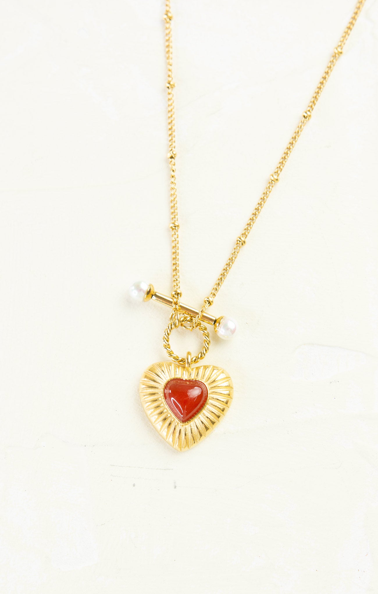 Gold necklace with a heart-shaped red pendant on a white background