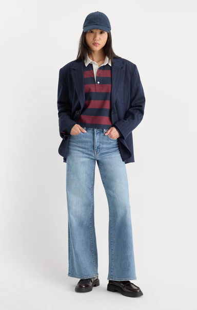 A model stands against a light gray studio background wearing a navy blazer, a navy cap, a navy and burgundy striped polo shirt, light-wash blue flare jeans, and dark brown loafers.