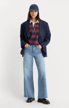 A model stands against a light gray studio background wearing a navy blazer, a navy cap, a navy and burgundy striped polo shirt, light-wash blue flare jeans, and dark brown loafers.