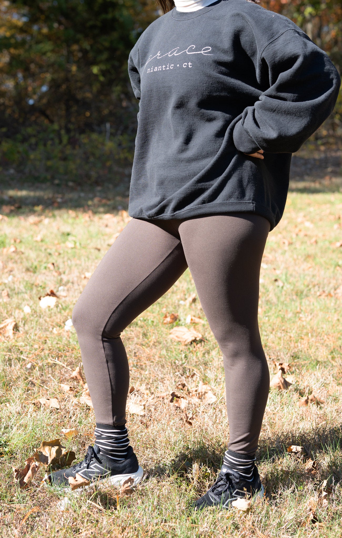 Person wearing a dark gray sweatshirt with text, dark leggings, and sneakers outdoors.