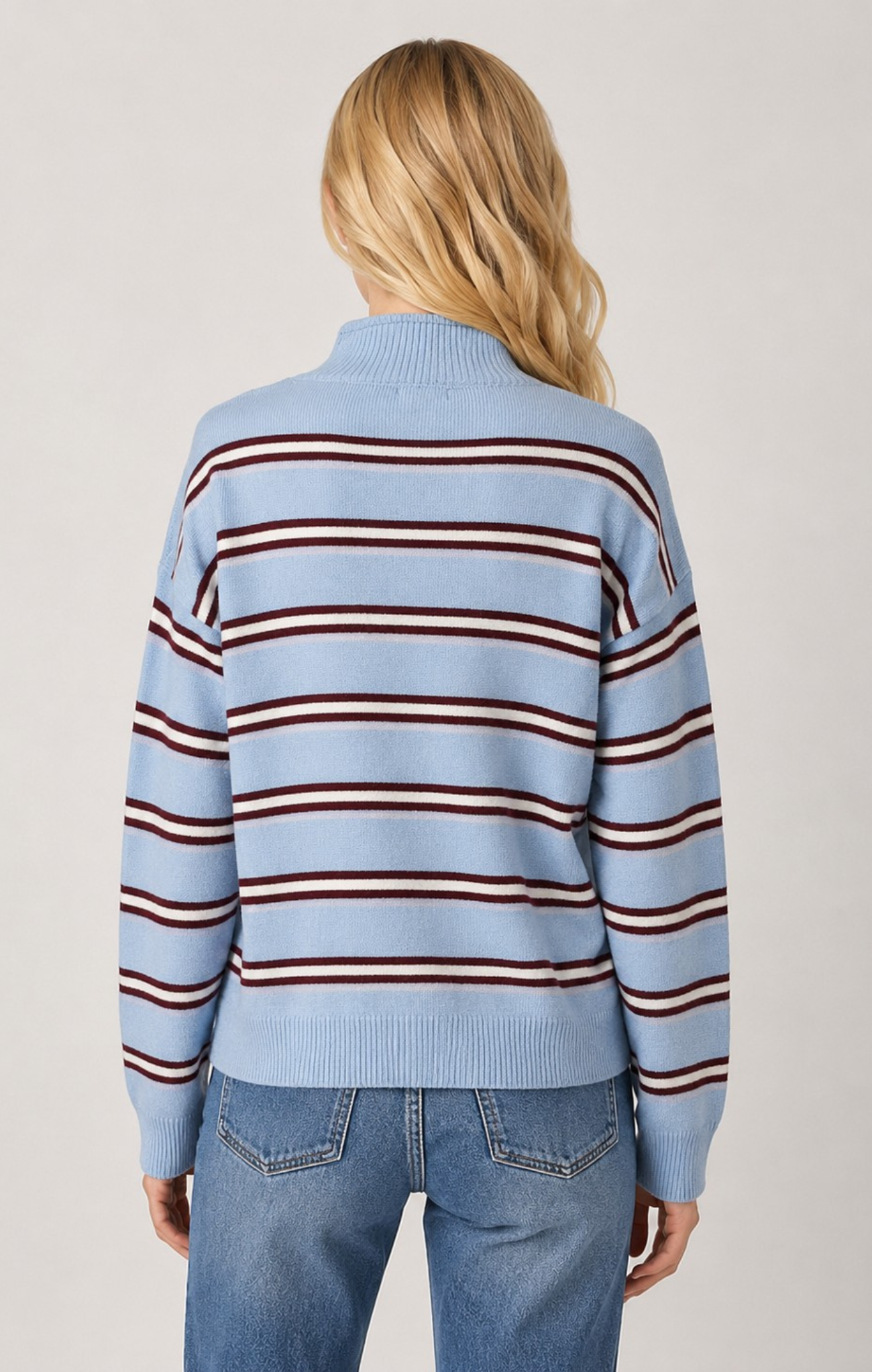 A woman viewed from the back wearing a light blue knit sweater with horizontal dark red and brown stripes, paired with blue jeans, against a plain light gray studio background.