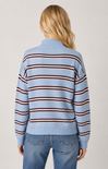 A woman viewed from the back wearing a light blue knit sweater with horizontal dark red and brown stripes, paired with blue jeans, against a plain light gray studio background.