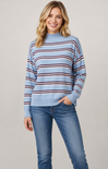 A woman wearing a light blue mock-neck sweater with horizontal navy and burgundy stripes, paired with blue jeans, against a solid light gray studio background.