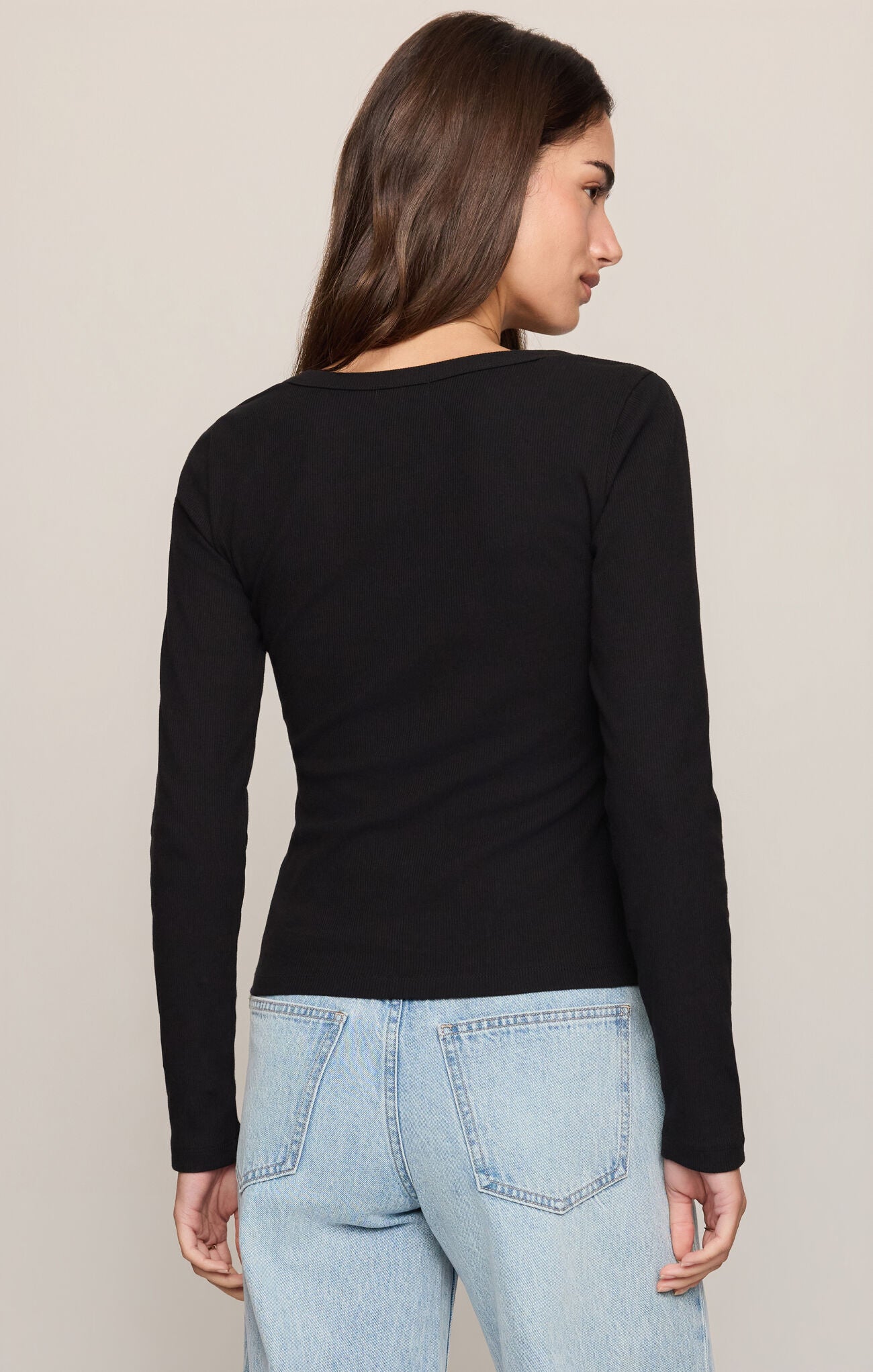 A woman shown from the back wearing a fitted black long-sleeve top and light blue jeans against a plain light beige studio background.