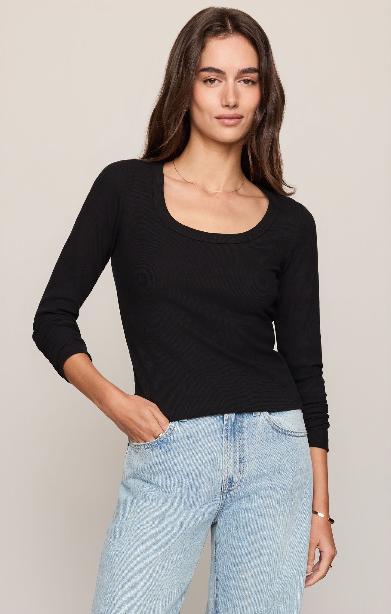 A woman modeling a black long-sleeve scoop-neck top paired with light-wash blue jeans against a plain beige studio background.