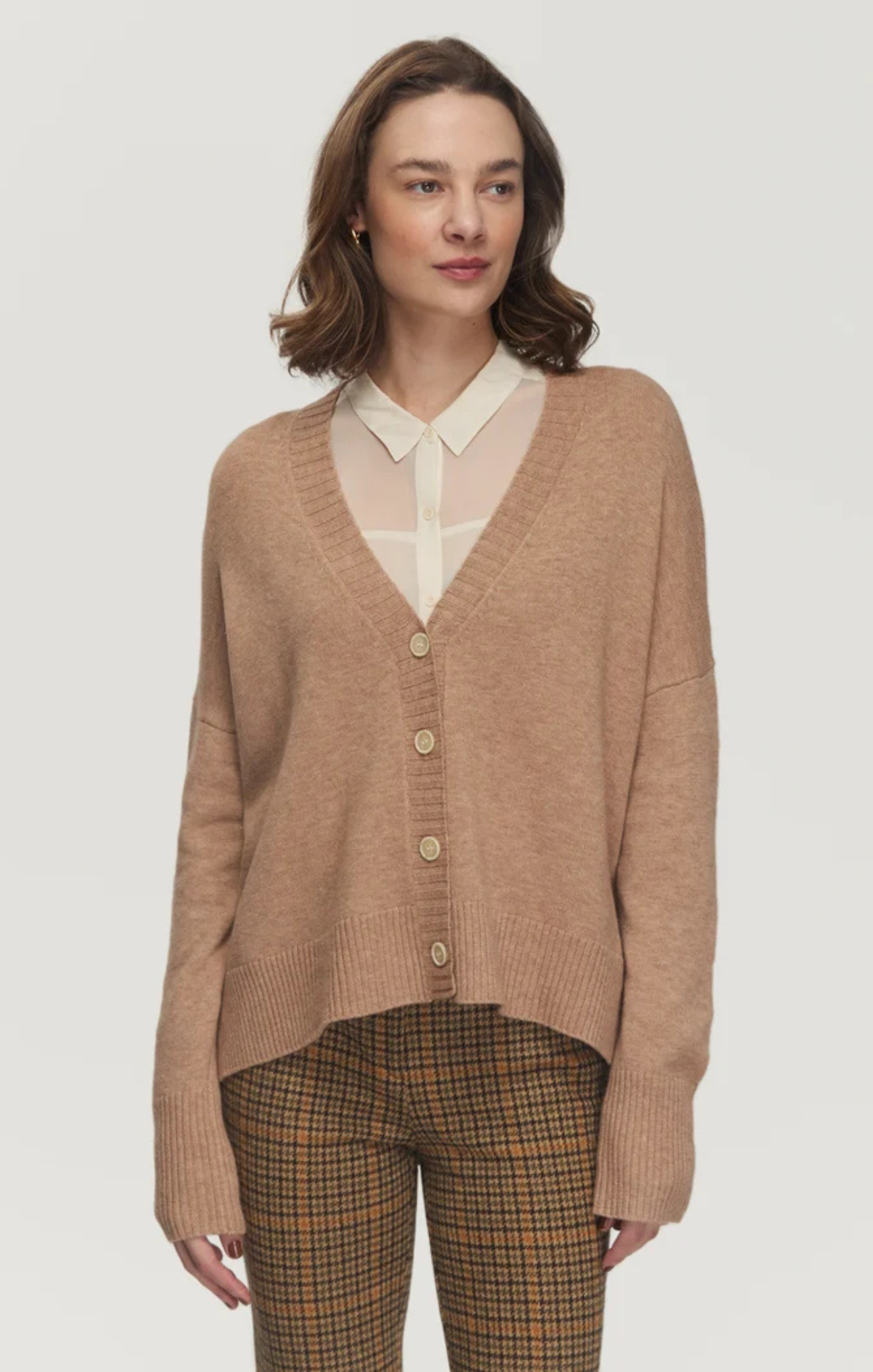 A woman modeling a light brown knit cardigan sweater over a cream collared shirt, paired with brown and beige plaid trousers, against a solid light gray background.