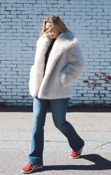 Person wearing a large white fur coat against a light blue brick wall.