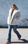 Person wearing a large white fur coat against a light blue brick wall.