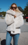 Woman wearing a fluffy white coat and sunglasses outdoors on a sunny day.