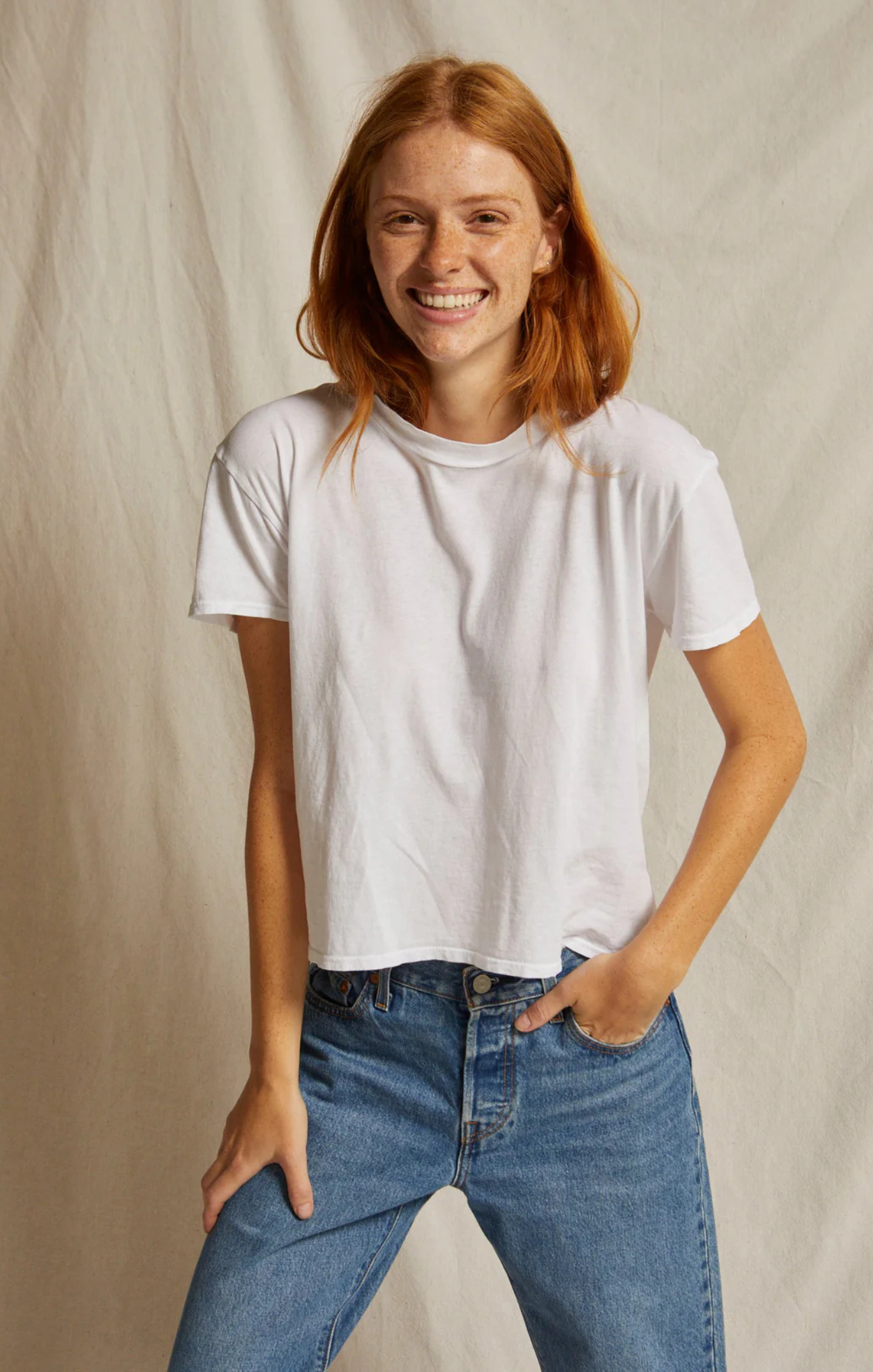 A smiling woman with red hair wearing a plain white short-sleeve crewneck t-shirt and blue jeans, standing against a neutral beige fabric backdrop.