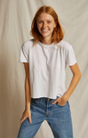A smiling woman with red hair wearing a plain white short-sleeve crewneck t-shirt and blue jeans, standing against a neutral beige fabric backdrop.
