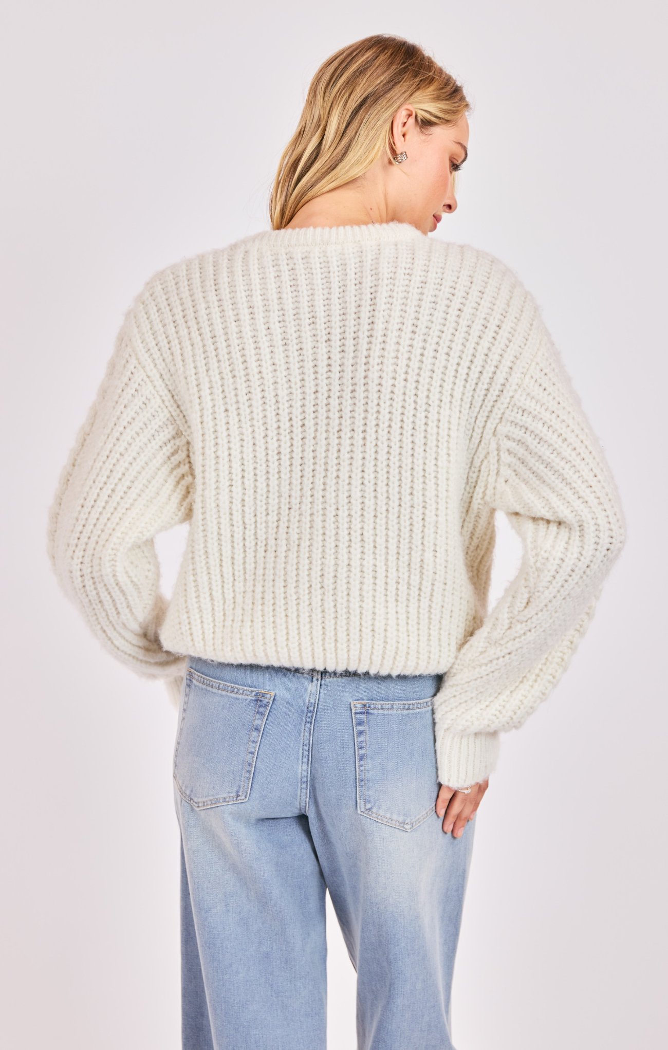 Woman wearing a cream chunky knit sweater and light blue jeans on a plain background