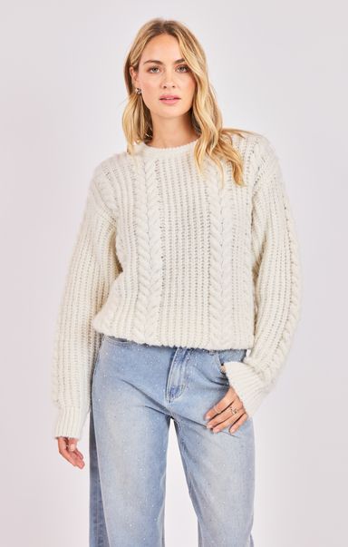 Woman wearing a cream chunky knit sweater and light blue jeans on a plain background