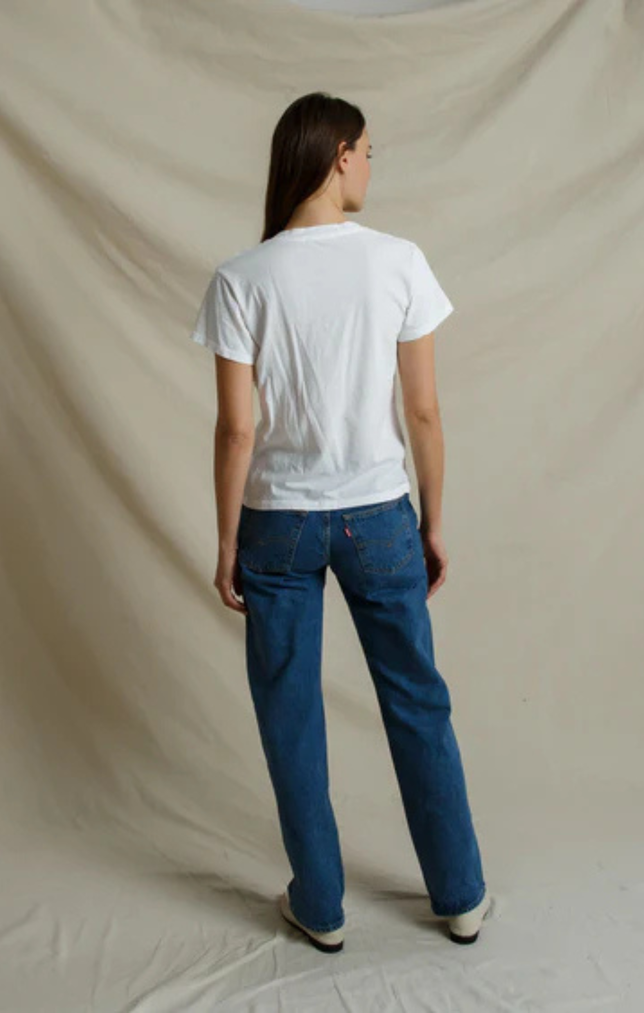 A model shown from the back wearing a white short-sleeve t-shirt and blue jeans, standing against a draped beige fabric backdrop.