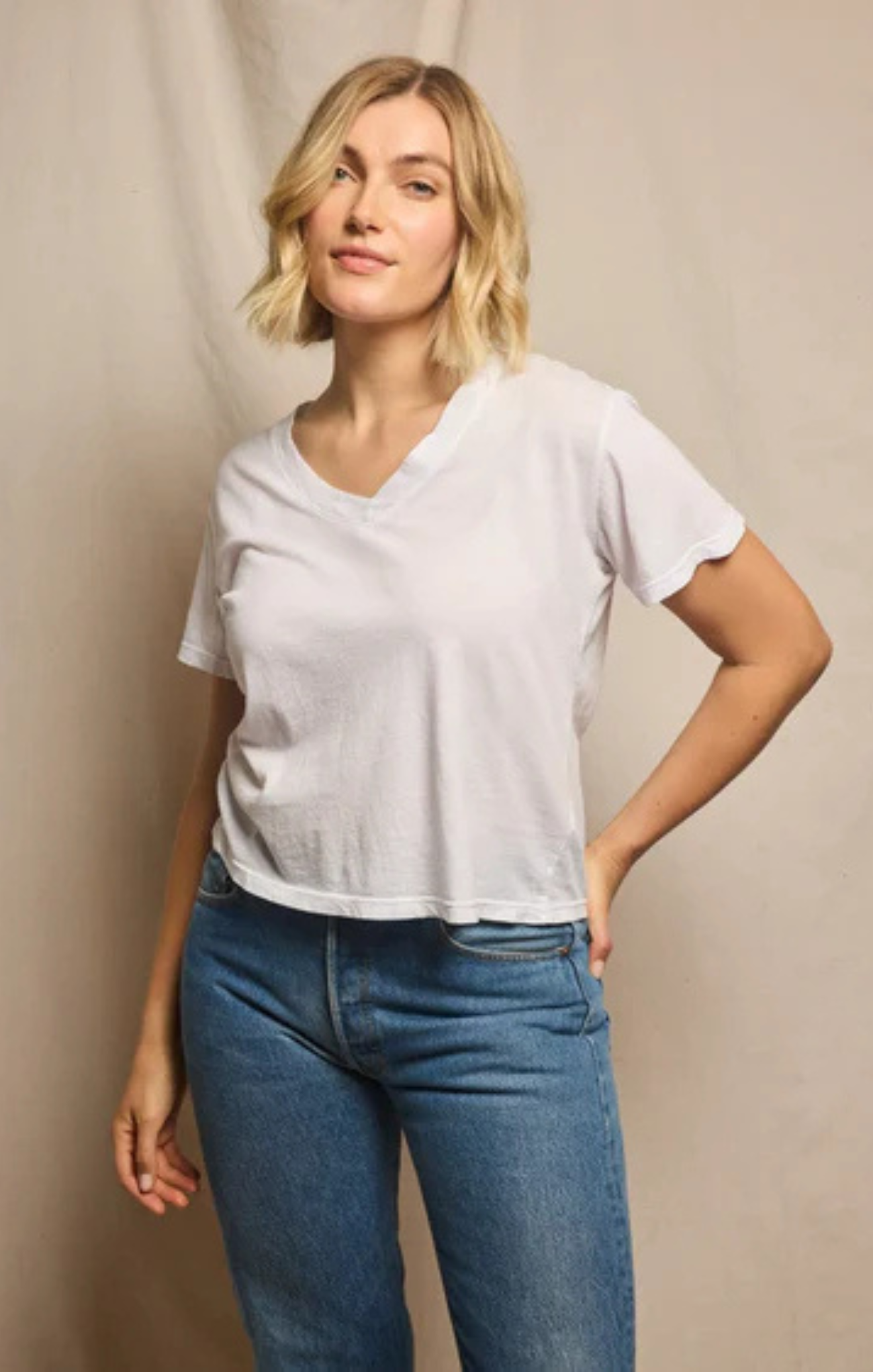 A woman modeling a light gray short-sleeve V-neck t-shirt paired with blue jeans, standing against a neutral beige backdrop.